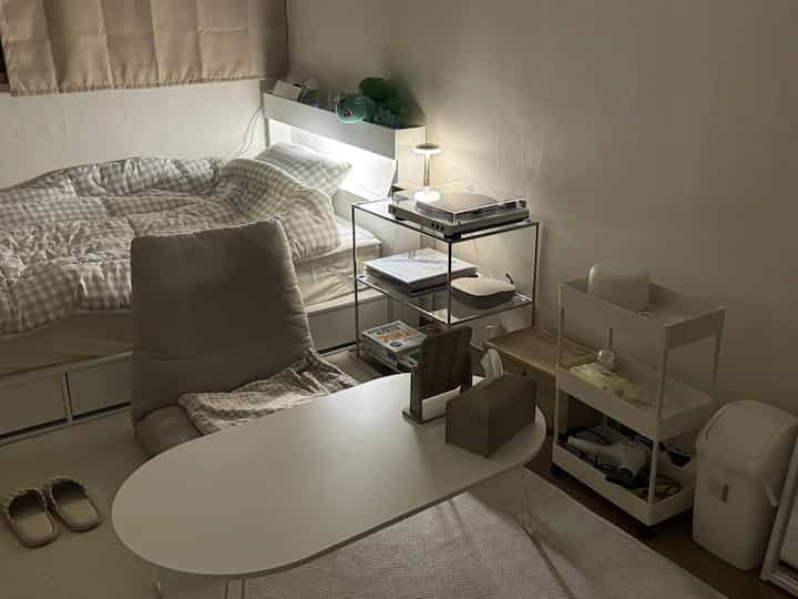 White and beige toned studio bedroom featuring a minimalistic bed, chair, and storage racks with a cozy and practical atmosphere