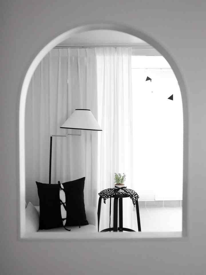 A white and monotone arch-shaped space featuring a white sofa, floor lamp, black cushion, and stool creating a simple living room interior