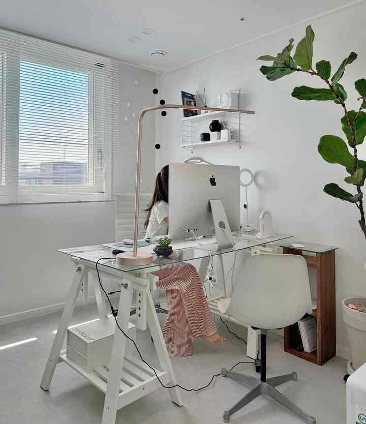 白色調のリビングルームのホームオフィス、ガラスの机と白いオフィスチェア、大きな植物とブラインドが配置された清潔感のある空間