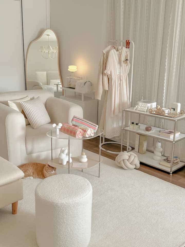 A cozy white and natural-toned studio bedroom featuring a sofa, curvy full-length mirror, clothing rack, and lace curtains