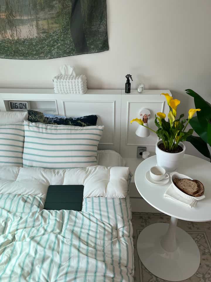 White and green toned bedroom featuring a bed with striped duvet cover centrally, and a round table with yellow flowers beside for cozy atmosphere