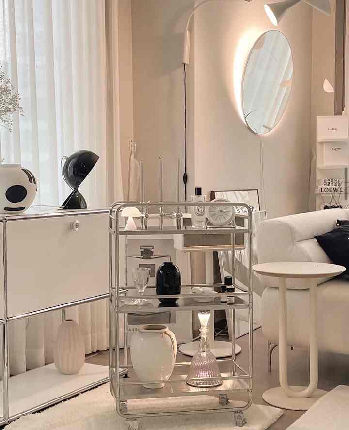 白とシルバーのトーンでまとめられた、モダンでミニマルなリビング。メタルトロリーと丸い鏡、白いソファが配置された印象的な空間