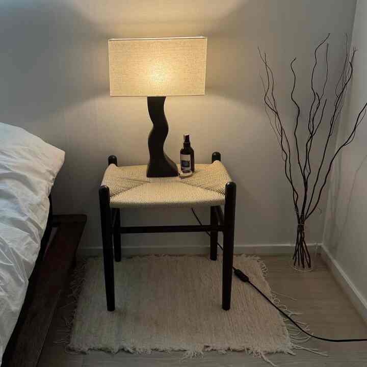 ナチュラルな色調の寝室、濃い木製のベッドフレームと編まれたスツールに置かれた照明がある雰囲気の良いインテリア