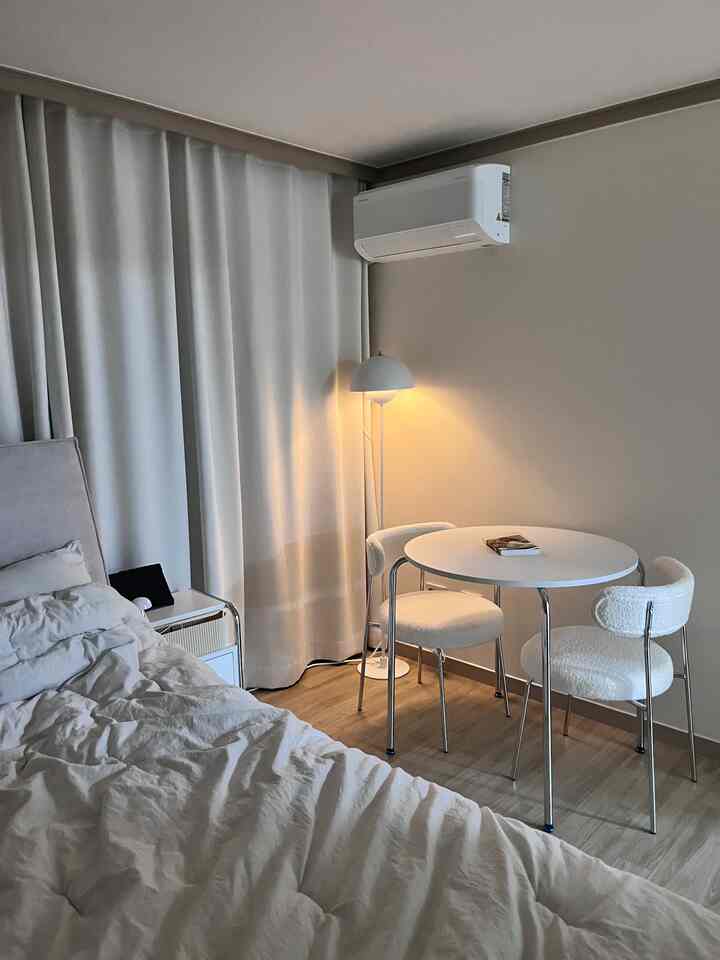 ホワイトとベージュのトーンによるシンプルモダンなリビング、丸いダイニングテーブルと2脚の椅子、快適なベッドが配置された落ち着いた空間