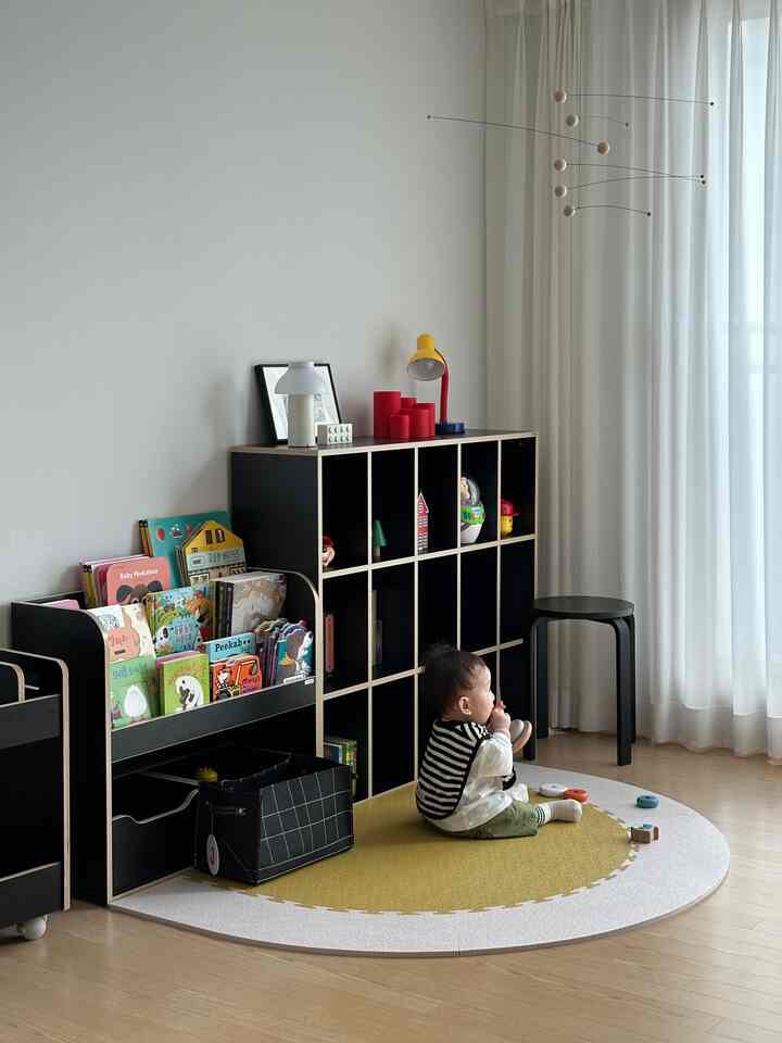 薄い木目調の床と白いカーテンがある子供部屋、黒い本棚と黄色のハーフムーンマットの上に赤ちゃんが座ったモダンで自然な雰囲気のインテリア
