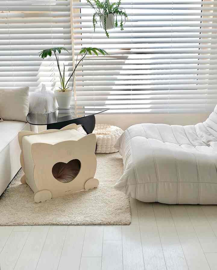 A white and beige toned living room featuring a sofa, light gray chaise lounge, and a wooden pet house near window blinds, creating a warm cozy atmosphere