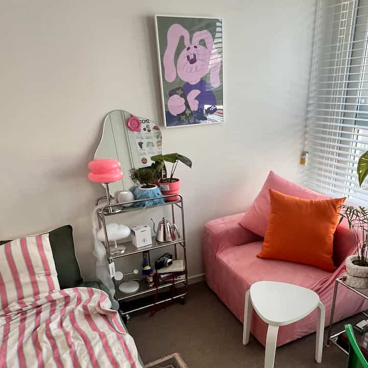 Soft pink and white toned living room featuring a pink sofa, white triangular stool, stainless steel trolley, and plants with a clean Mid-Century Modern style