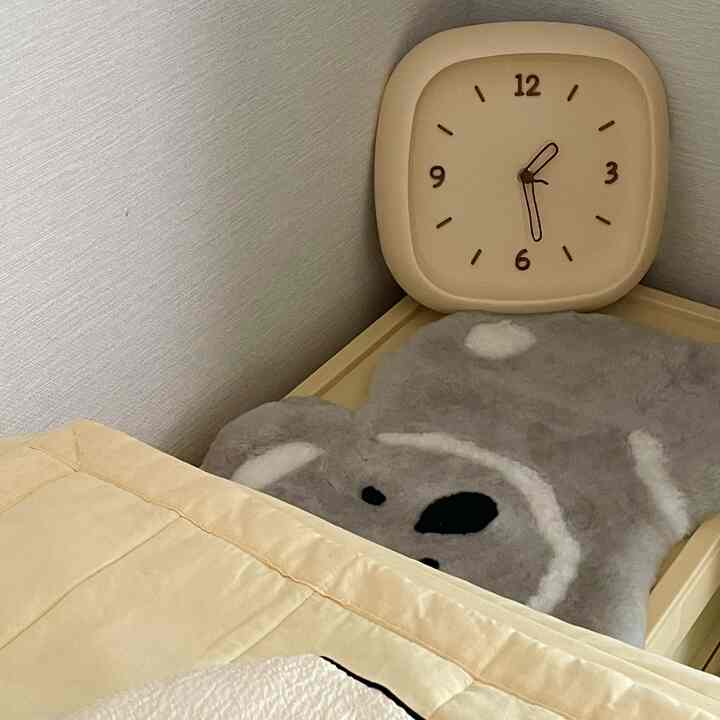 柔らかなベージュと木目調の調和が感じられる6畳の寝室、ナイトテーブルの上に置かれた味わい深い時計とかわいいクマのマットが配置された温かみのある空間