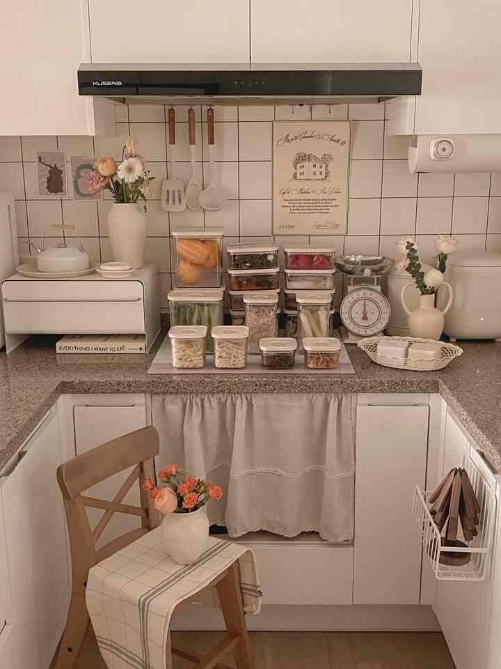 アイボリーとベージュの色調のキッチン、整然と並んだ密閉容器と木製の椅子、花瓶が配置された雰囲気のある空間