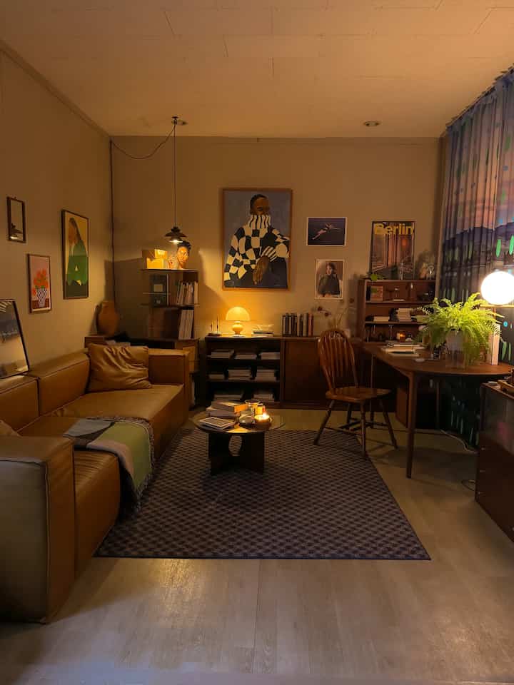 温かみのある木目調とベージュ色の壁が特徴のワンルーム書斎、レザーソファや机、本棚、多様な照明が配置された雰囲気のあるインテリア