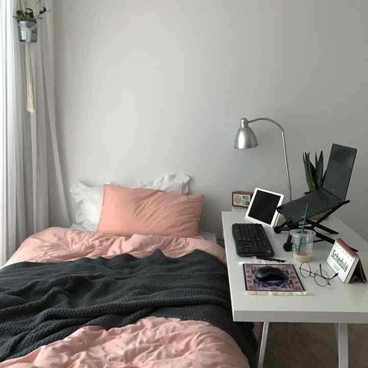 白い壁とカーテンが調和する寝室、ピンクの寝具とグレーのスロー、ノートパソコンを置いたデスクが配されたシンプルなホームオフィス兼寝室