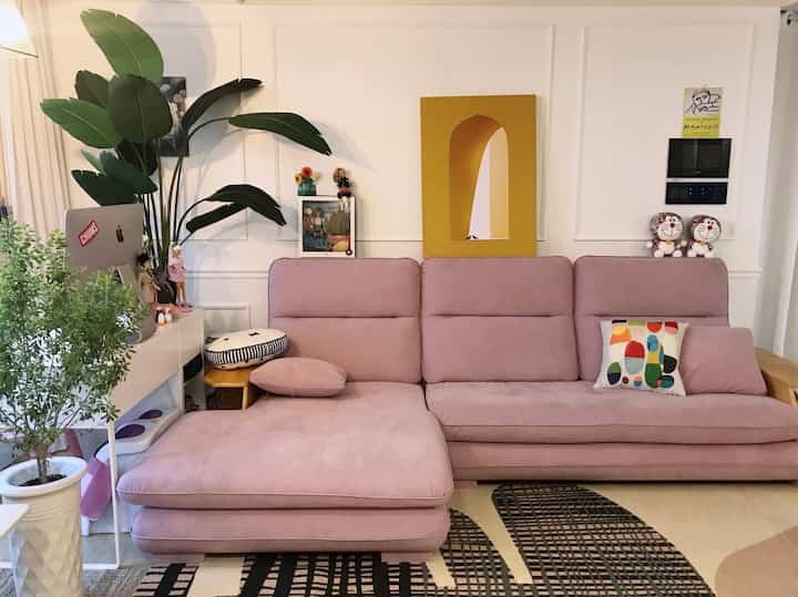 Living room with white walls and wainscoting, featuring a pink sectional sofa, plants, and cushions creating cozy atmosphere
