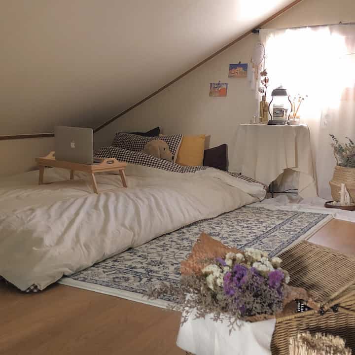 Soft natural-toned loft bedroom with low slanted ceiling, cozy bedding, candles, and a rug creating a warm atmosphere