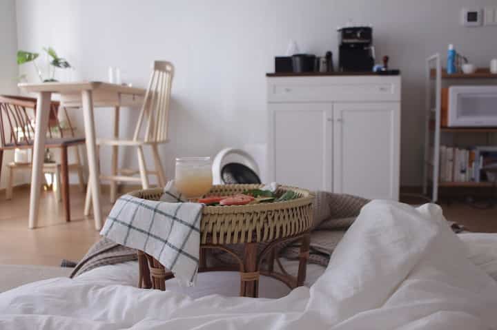 A white and natural wood tone studio bedroom featuring a dining table and a home cafe tray on the bed, creating a cozy atmosphere