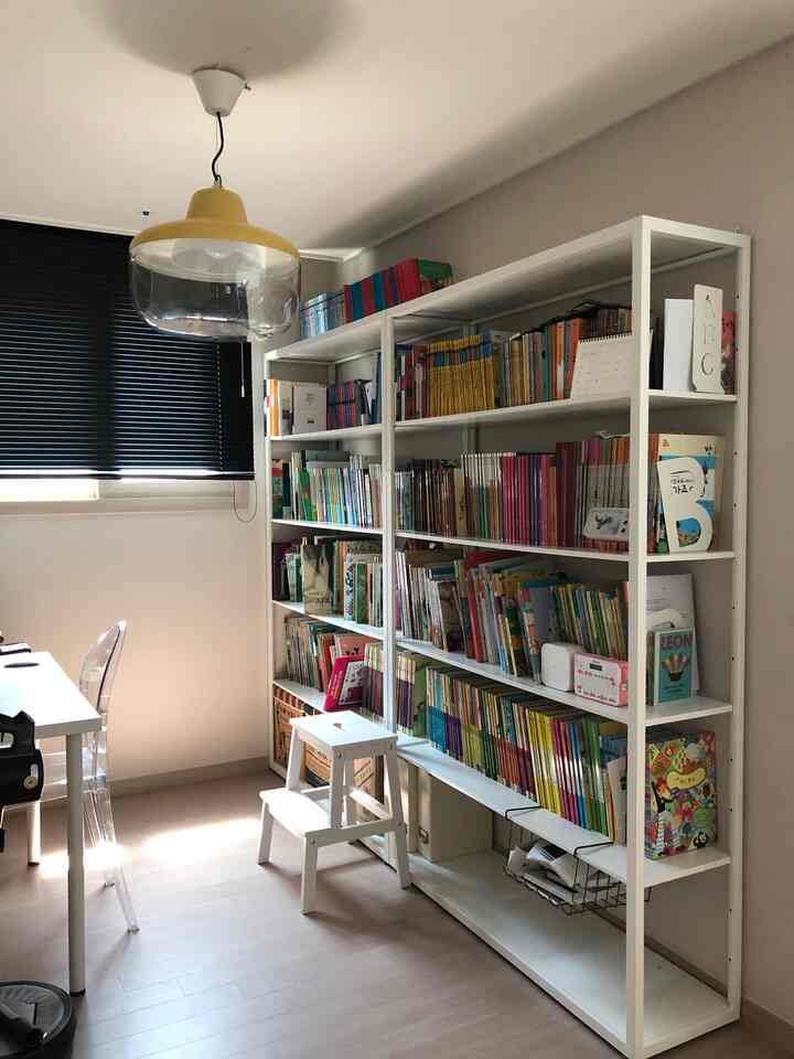 White-dominant bedroom with bookshelf and stool under a black window blind and yellow pendant light