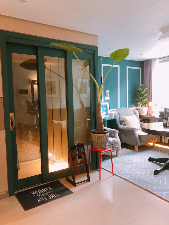 緑色の壁と網入りガラスの引き戸があるナチュラルモダンな玄関とリビング空間、大きな植物とグレーのアームチェアが配置された落ち着いた雰囲気のインテリア