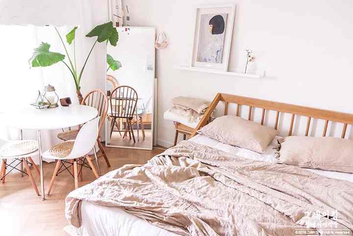 A bright bedroom in white and beige tones featuring a wooden bed frame, round dining table, and full-length mirror in a natural and simple setting