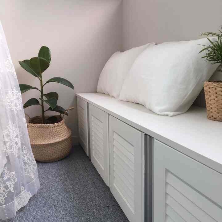 Cozy balcony space featuring gray carpet, white bench with pillows, and potted plants in natural baskets