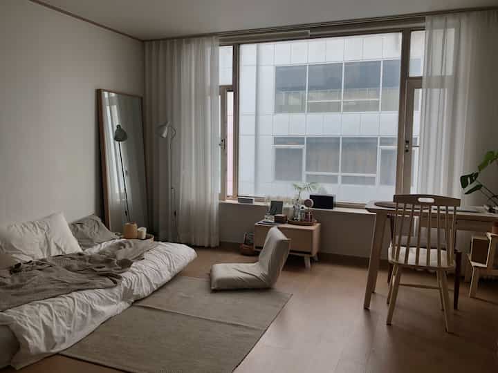 A cozy and tidy studio apartment in beige and natural tones featuring a mattress bed, desk, and floor lamp