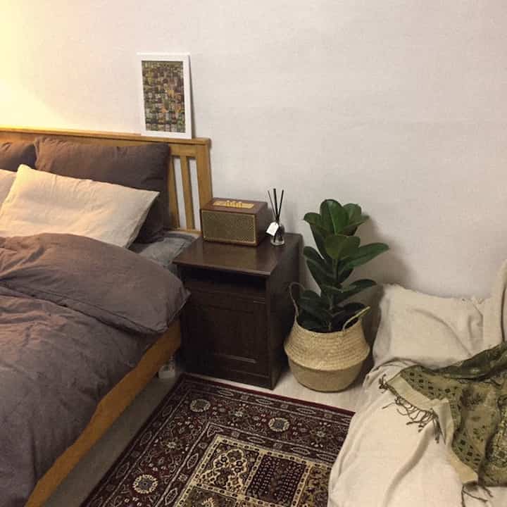 Natural wood and beige toned bedroom space featuring a nightstand with a Bluetooth speaker and a plant, creating a cozy atmosphere