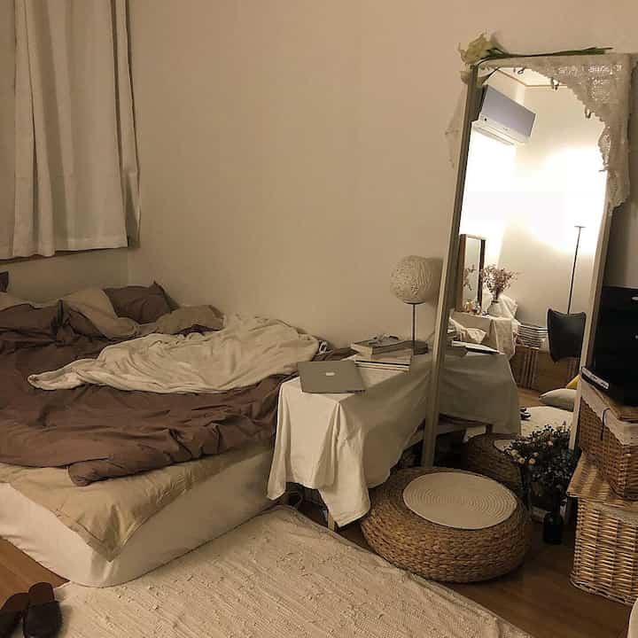 Beige-toned studio bedroom featuring a natural woven stool and full-length mirror with warm, cozy ambiance