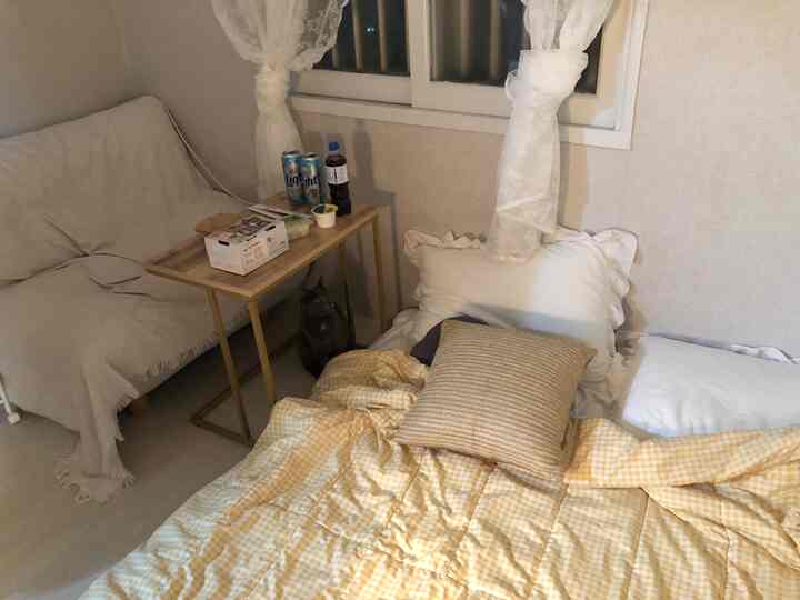White and beige toned single household bedroom-living room featuring sofa, side table, and lace curtains creating cozy atmosphere