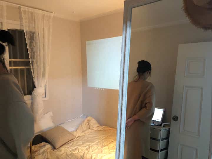 Beige-toned studio bedroom featuring a projector screen on the wall, bed, and a mobile storage trolley in a cozy setting