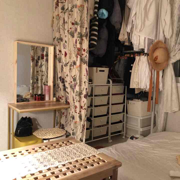 ホワイトとウッドトーンが調和した狭い寝室。花柄カーテンで隠れたハンガーと木製の化粧台が配置された雰囲気のあるインテリア