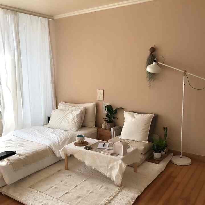 Natural tone bedroom with beige walls and white curtains featuring low beds, floor seating with cushions, and indoor plants in a cozy setting