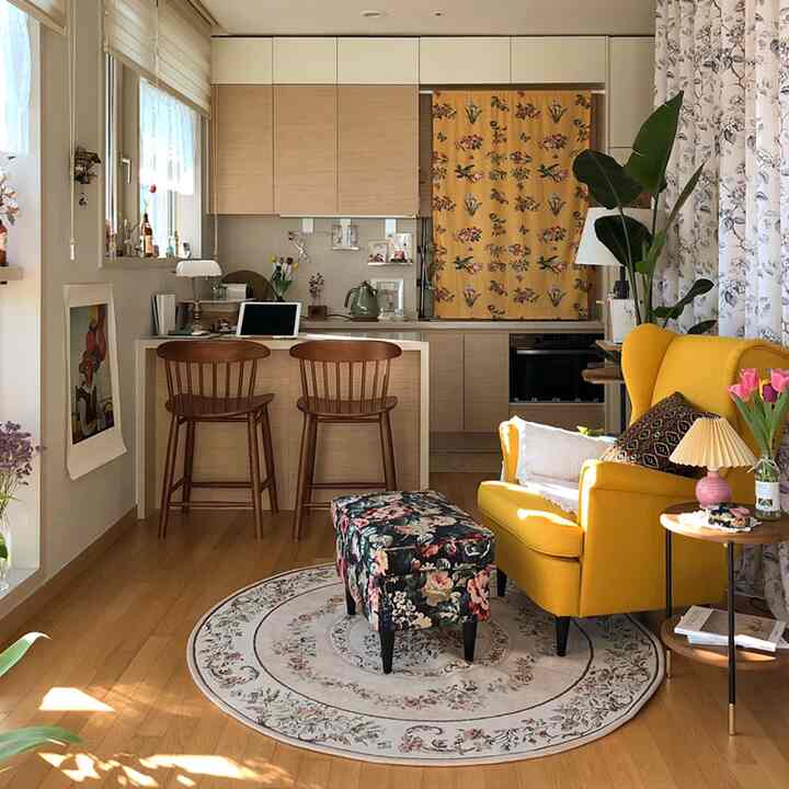 Warm-toned studio apartment living area with bright yellow armchair and floral patterned ottoman near wood-tone kitchen