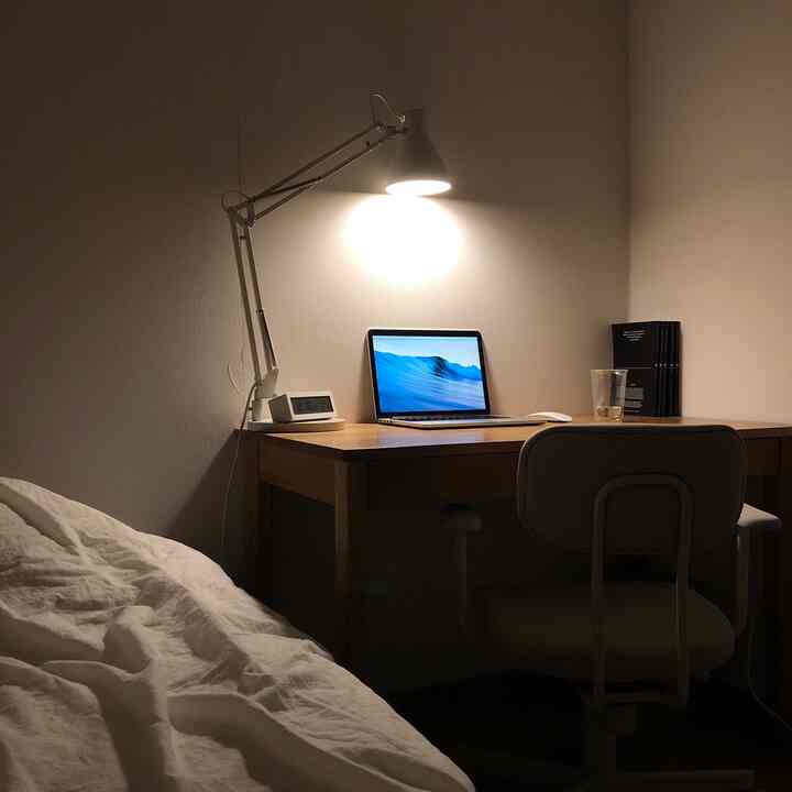 暖かい照明が灯る木製デスクとノートパソコンが置かれた一人暮らし向けの小さな寝室兼ホームオフィスの空間
