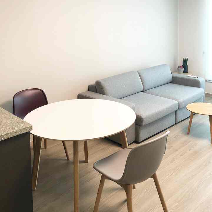 Modern living room with white walls and natural wood-tone flooring, featuring a gray sofa and round dining table in a simple setting