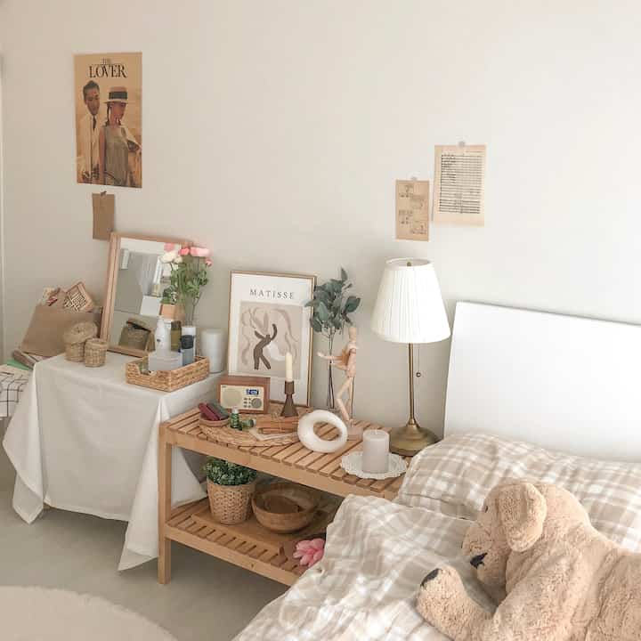 ベージュと白を基調としたワンルーム寝室、ベッドと木製棚、照明と額装のある自然でシンプルな印象のインテリア