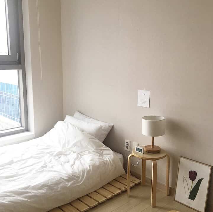 Minimal bedroom with beige walls and wood-toned decor, featuring a low wooden bed and a round stool with a table lamp