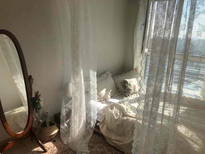 A white and beige toned bedroom featuring a natural wood mirror and lace curtains with a warm atmosphere.