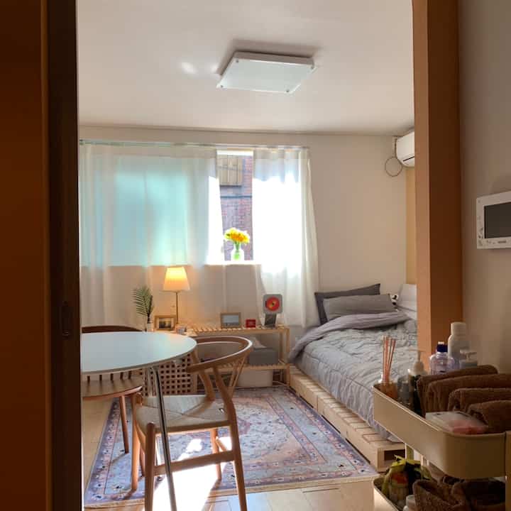 明るいホワイトとナチュラルウッド調のワンルーム寝室、丸いダイニングテーブルとラグが配置された雰囲気のあるインテリア