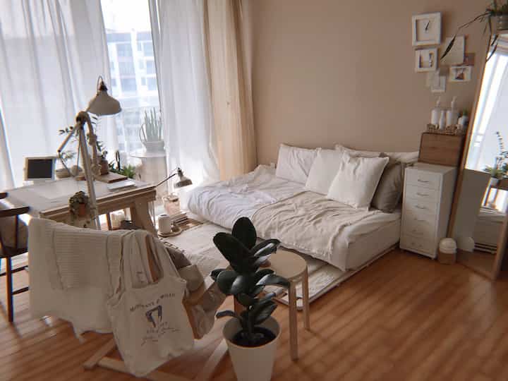 Beige and white toned bedroom featuring floor mattress, wooden desk, and plants in a natural setting