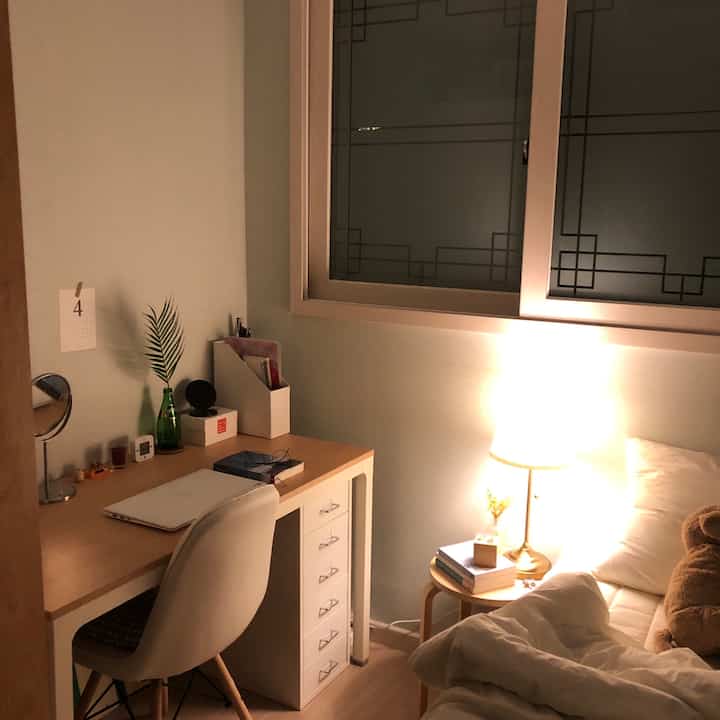 白とナチュラルカラーのワンルーム寝室、机とベッド、温かみのあるテーブルランプが配置された雰囲気のあるインテリア