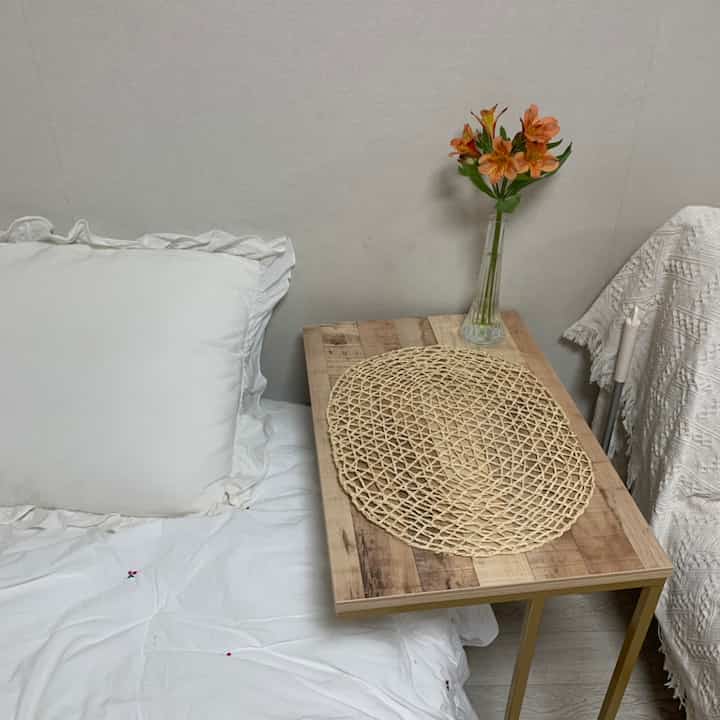 Natural tone bedroom space featuring a wooden side table, white bedding, and a vase with flowers creating a cozy atmosphere