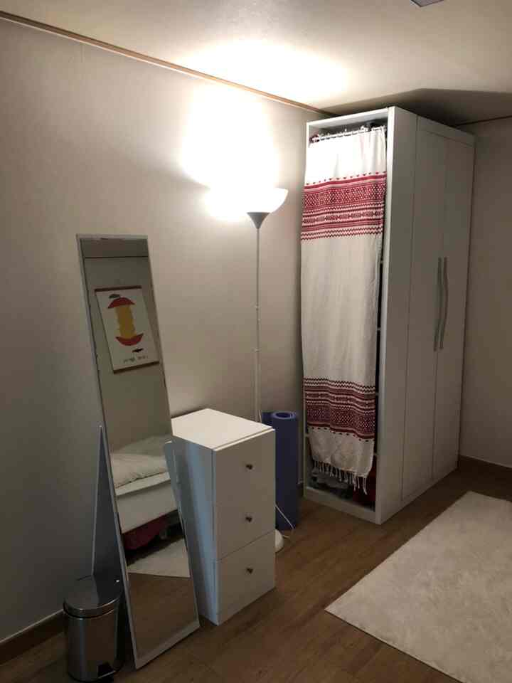 白と木目調が調和したワンルーム寝室、ワードローブ、化粧台、立て掛け鏡、フロアランプが配置された落ち着いた空間