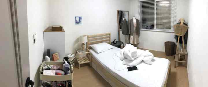 Bright natural-toned small bedroom featuring a wooden bed, nightstand, full-length mirror, stool, and clothing rack by the window