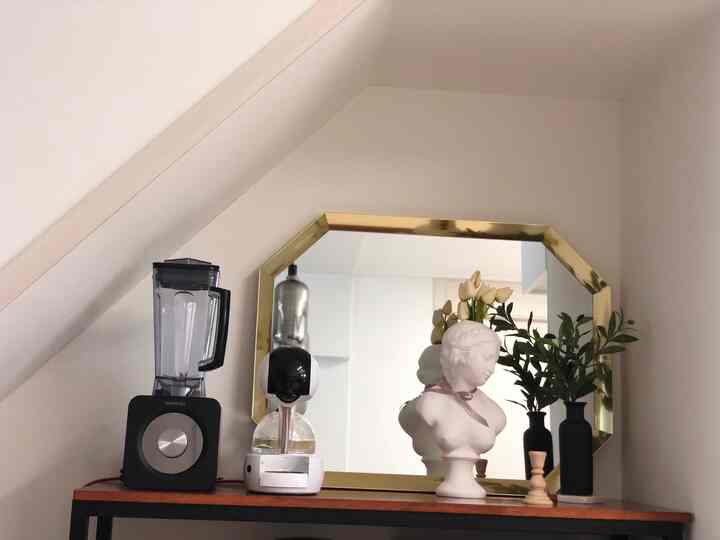 白い壁とゴールドの鏡枠がある階段下のスペース、棚の上に置かれた彫刻とブレンダーが配置された印象的なおうちカフェコーナー