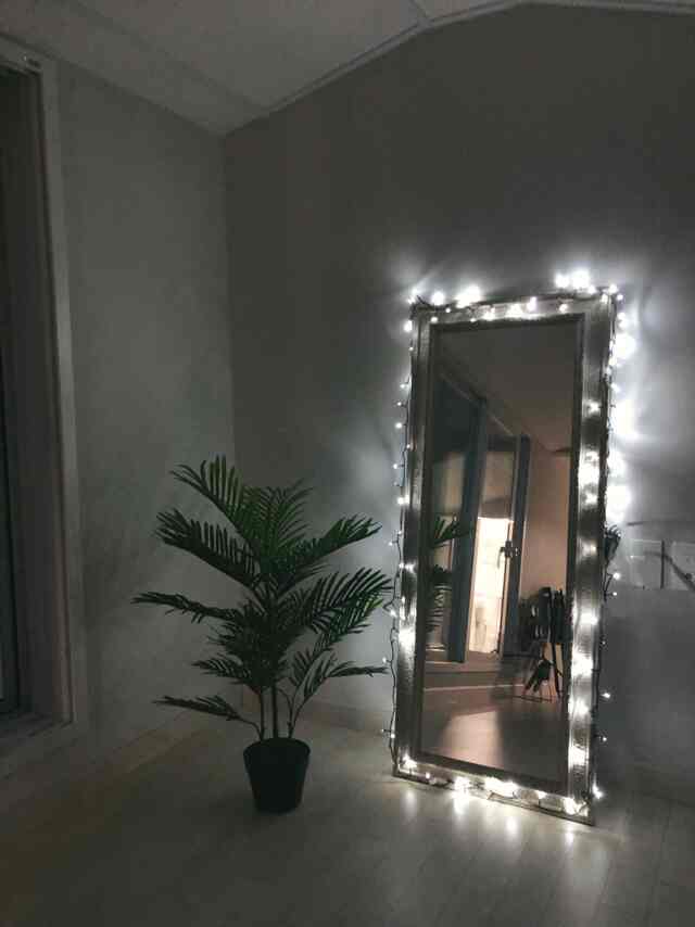 白い床とグレーの壁がある落ち着いたリビングのコーナー、人工のヤシの植物とストリングライトが巻かれた全身鏡が配置された空間