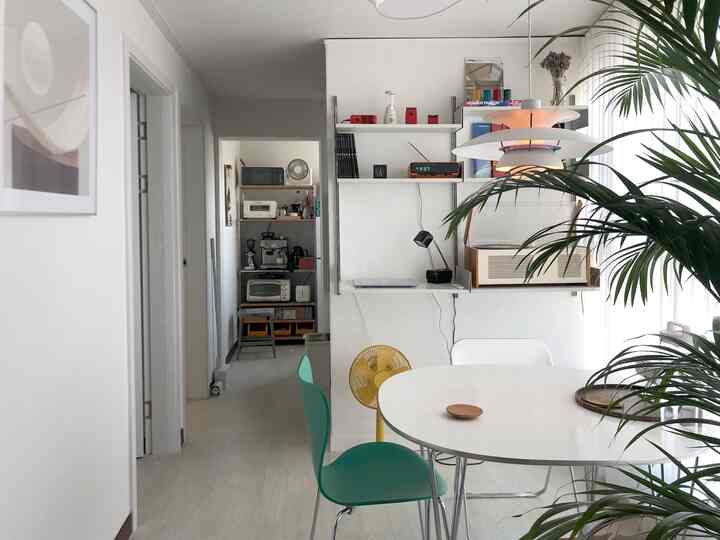 White and green toned studio apartment living room with round table, shelving system, and Louis Poulsen pendant light creating a cozy atmosphere
