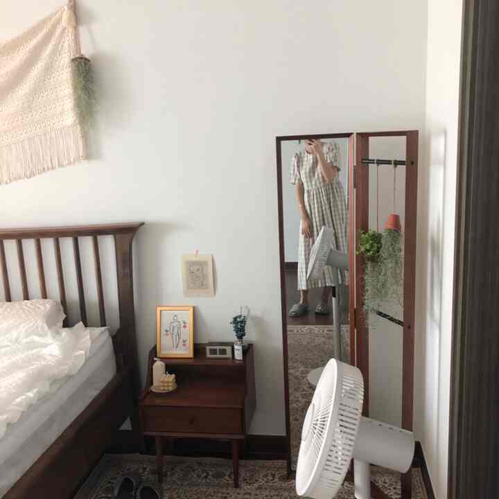Natural tone bedroom with white walls, brown wooden bed, full-length mirror, plants, and fans creating a calm atmosphere