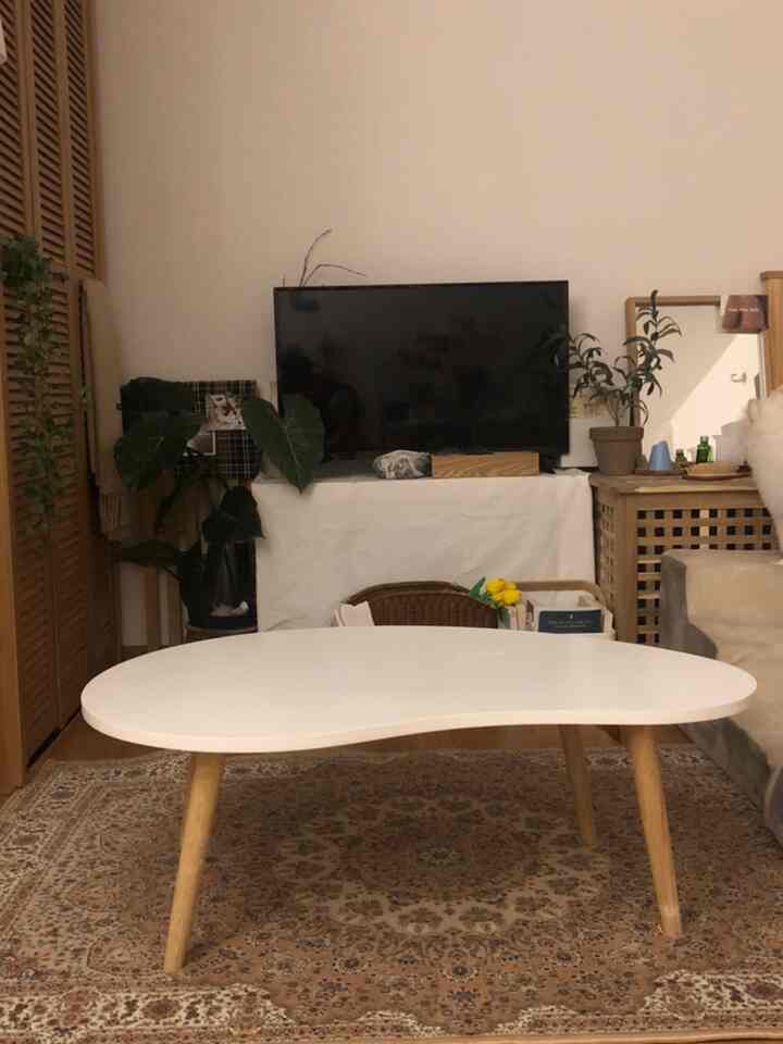 ナチュラルカラーのコンパクトなリビング、モダンな白いコーヒーテーブルと木製の脚、パターンラグ、テーブルクロスが掛けられたテレビ台が配置された雰囲気のある空間