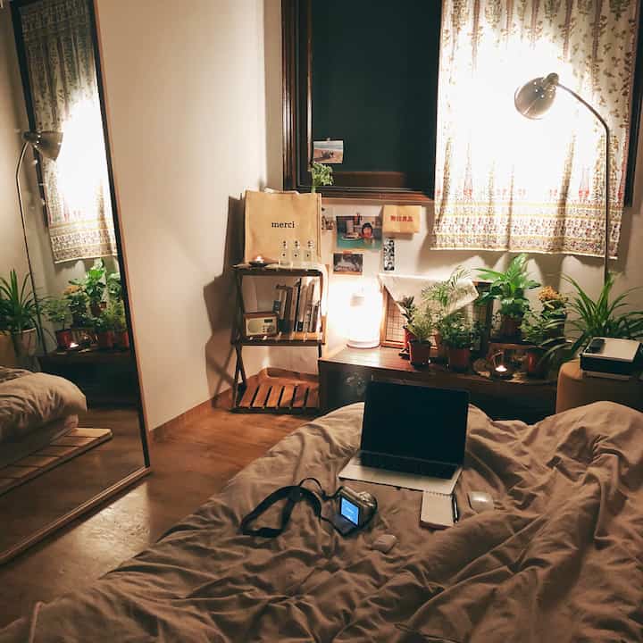 温かみのあるブラウンとベージュの寝室、ベッドにノートパソコンとカメラ、窓辺に植物と間接照明が配置された居心地の良いワンルーム