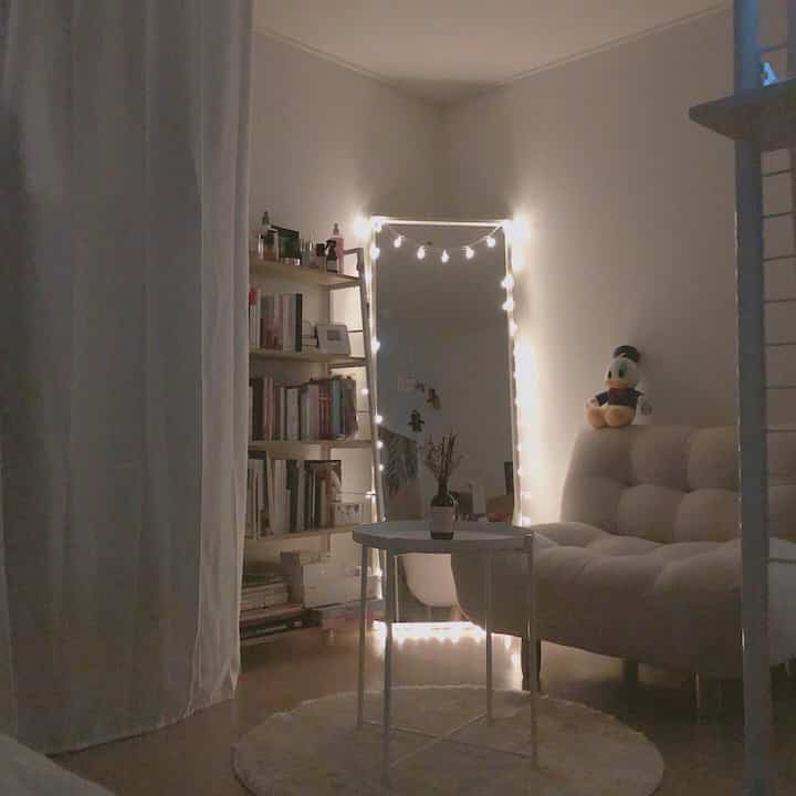 White-toned small room featuring white sofa, rug, and full-length mirror decorated with LED string lights, creating a cozy atmosphere