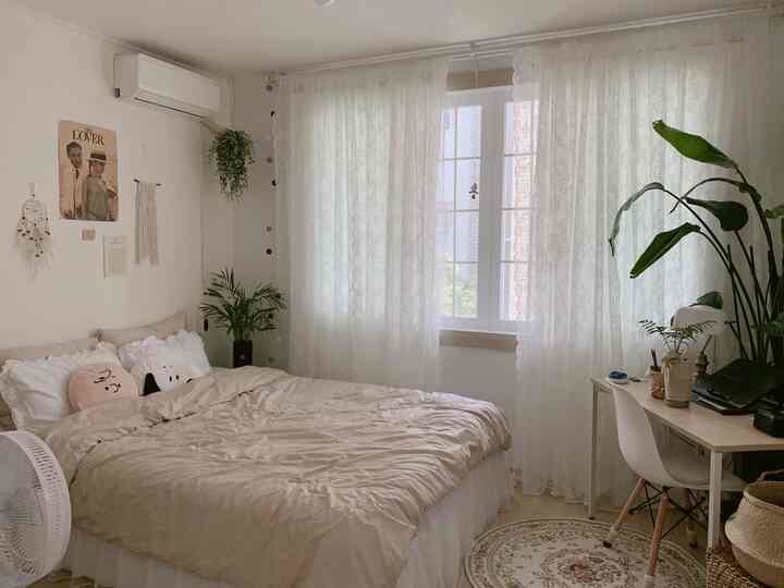 A white and beige toned bedroom featuring a bed, desk, and several plants, creating a natural and cozy atmosphere