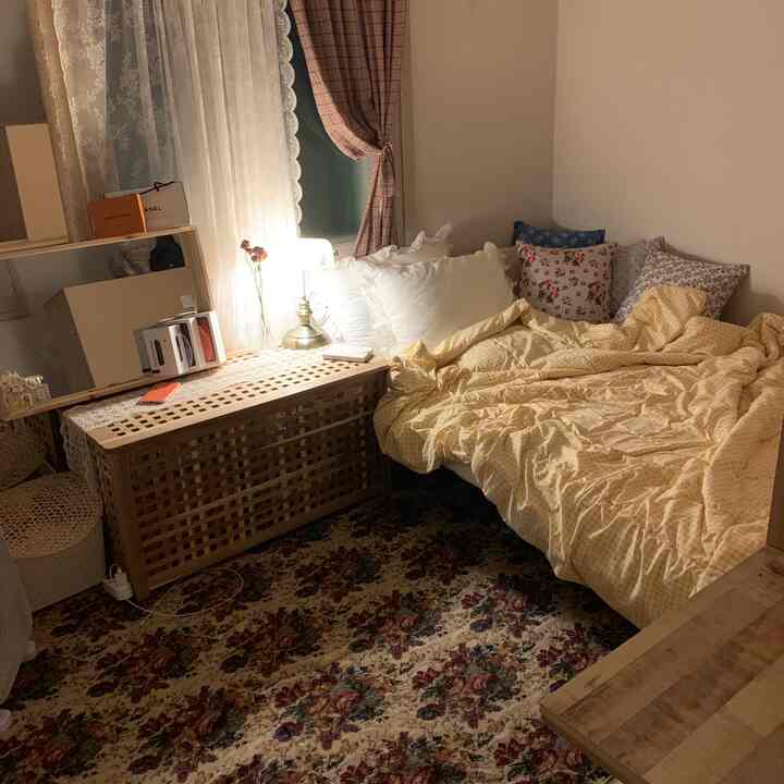 Cozy studio bedroom with yellow bedding and floral rug, natural wood storage table, lace curtains, and warm lighting
